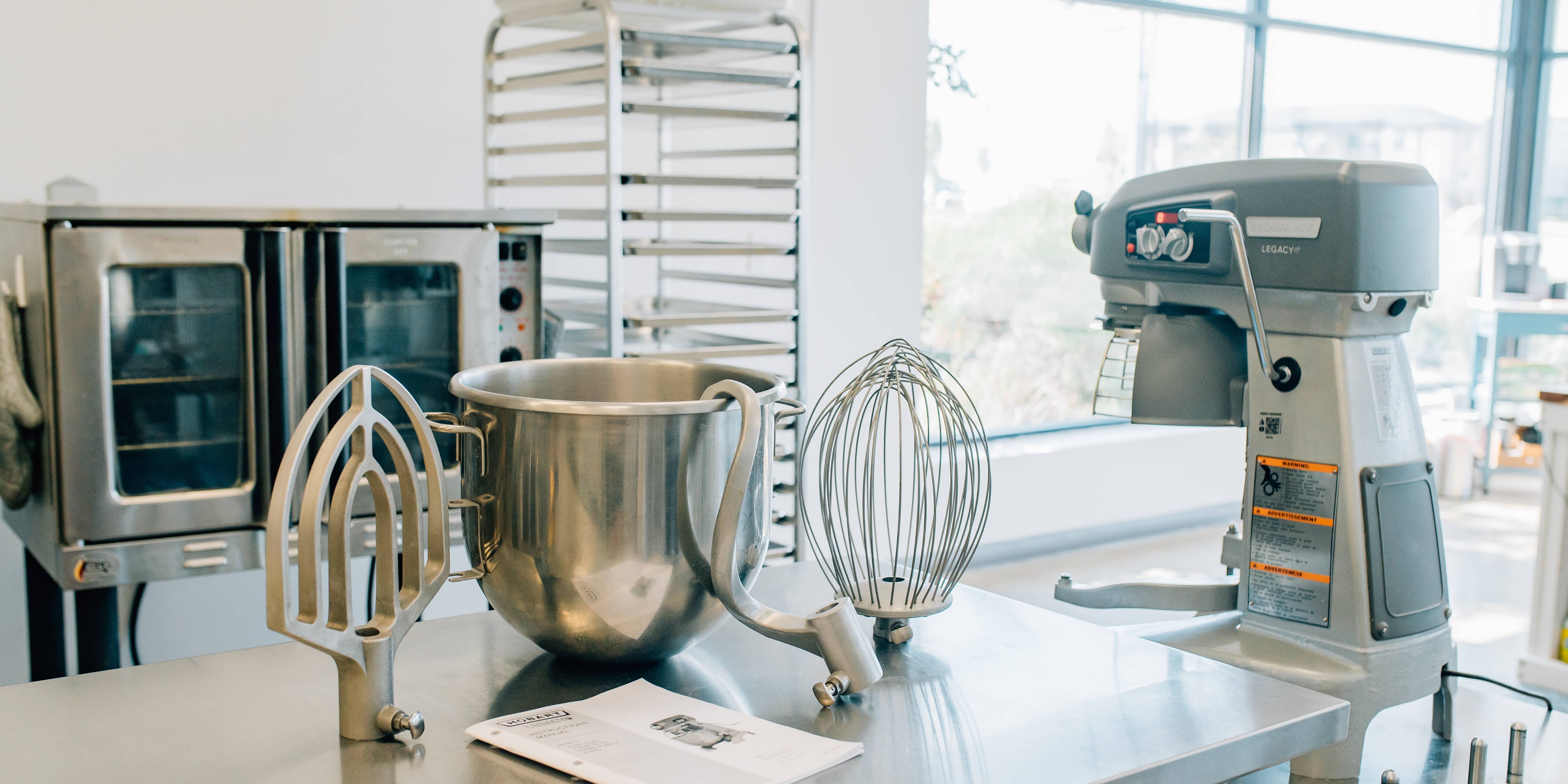 Beater, bowl, whip, and dough hook sit on a table to the left of a Hobart Legacy+ HL200 mixer viewed from the back
